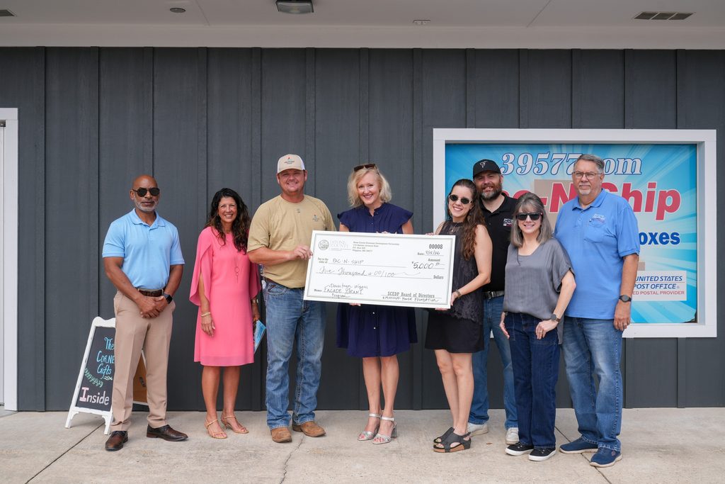 Eight people stand outside the Pac-N-Ship building in Wiggins, Mississippi, holding a ceremonial $5,000 check from the Stone County Economic Development Partnership and Mississippi Power Foundation, dated August 18, 2026, for the Downtown Wiggins Facade Grant Program.